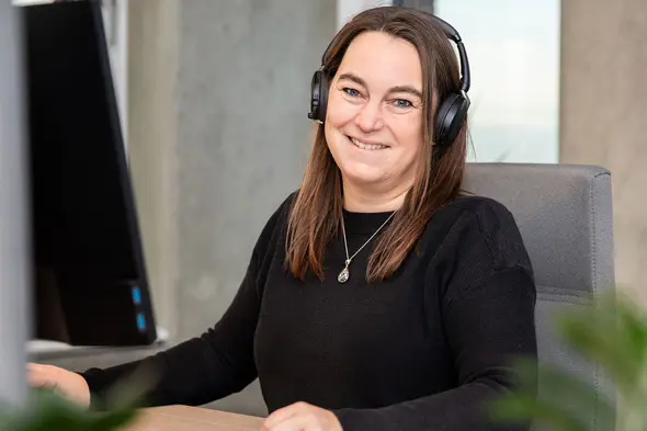 Femme souriante portant un casque audio, assise devant un ordinateur. Elle a les cheveux longs et bruns, et porte un pull noir. Arrière-plan flou avec des éléments modernes et un éclairage naturel.