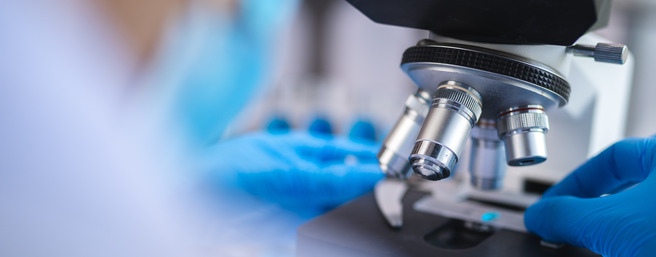 Un scientifique en blouse blanche utilise un microscope, avec une main tenant une lame. Des gants bleus sont visibles, et l'arrière-plan suggère un laboratoire. L'accent est mis sur le microscope et les outils scientifiques environnants.
