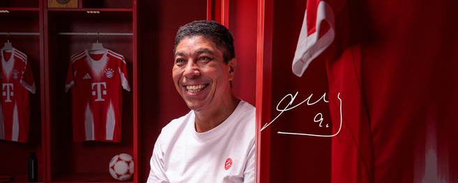 Un homme souriant est assis dans un vestiaire aux murs rouges, entouré de maillots de football. On peut voir un ballon de football et des éléments de l'équipe sur des étagères. À côté de lui, un dessin représentant sa signature est visible.