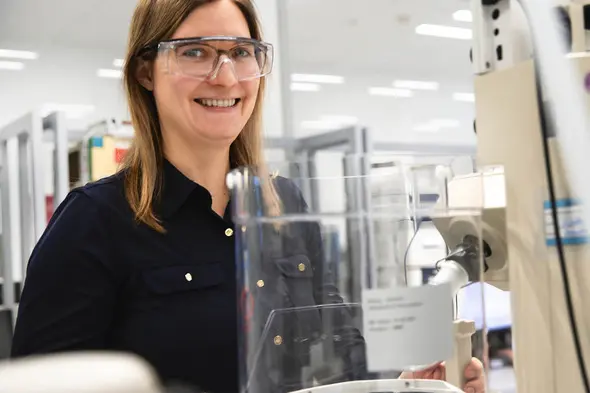 Femme souriante portant des lunettes de protection, se tenant près d'un équipement de laboratoire. L'environnement est moderne et bien éclairé, avec des étagères d'articles de laboratoire en arrière-plan.