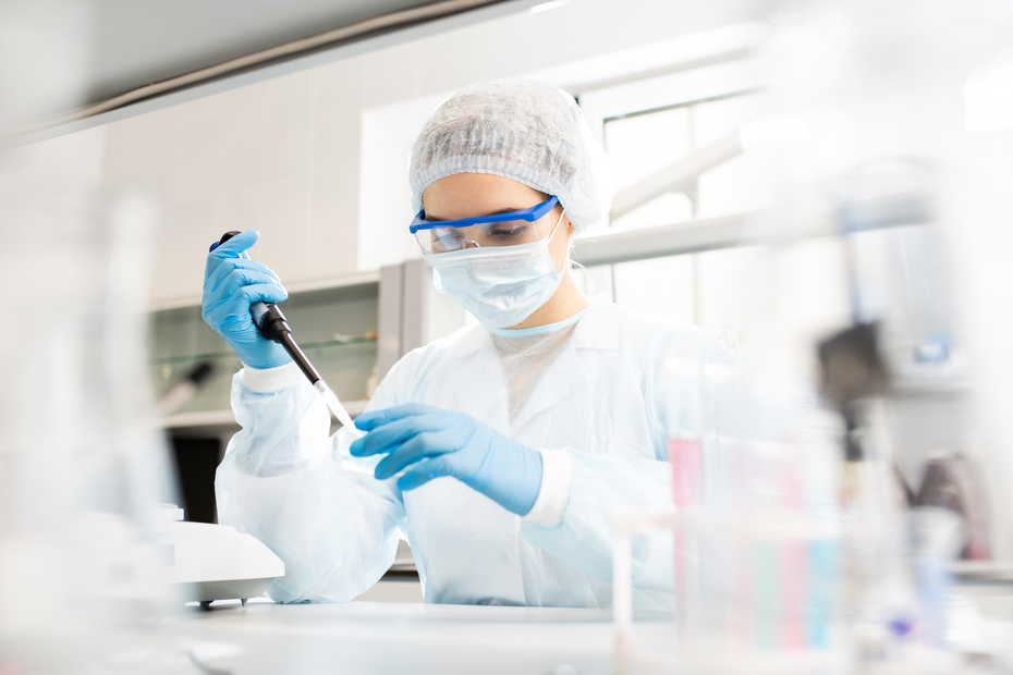 Une scientifique en blouse de laboratoire, portant un masque et des lunettes de protection, utilise une pipette pour manipuler un liquide. L'environnement est un laboratoire bien éclairé, avec des équipements de recherche en arrière-plan.