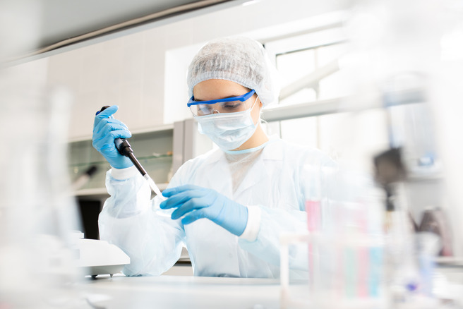 Une scientifique en blouse de laboratoire, portant un masque et des lunettes de protection, utilise une pipette pour manipuler un liquide. L'environnement est un laboratoire bien éclairé, avec des équipements de recherche en arrière-plan.