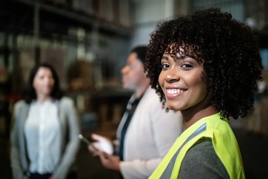 Femme souriante portant un gilet de sécurité contrasté, se tenant dans un entrepôt. Deux autres femmes se trouvent en arrière-plan, discutant. L'ambiance est celle d'un environnement professionnel.