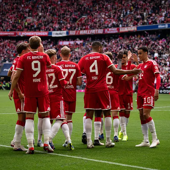 Un groupe de joueurs de football du FC Bayern Munich en tenue rouge se regroupe sur le terrain. Ils semblent discuter ou célébrer un moment ensemble, avec des supporters en arrière-plan.