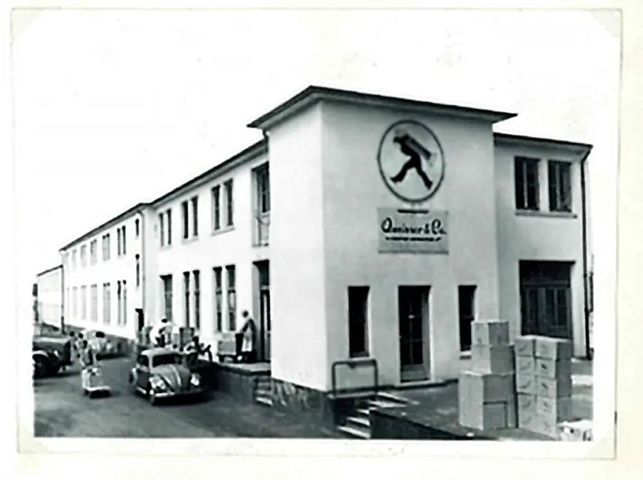 Bâtiment industriel en noir et blanc avec un logo au centre. Des personnes chargent des caisses à l'extérieur, et des voitures anciennes sont garées à proximité. L'architecture est simple, avec de grandes fenêtres et une entrée principale.