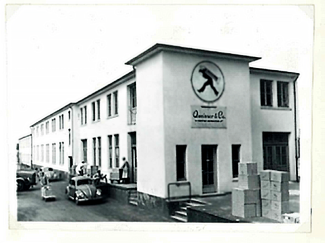 Bâtiment industriel en noir et blanc avec un logo au centre. Des personnes chargent des caisses à l'extérieur, et des voitures anciennes sont garées à proximité. L'architecture est simple, avec de grandes fenêtres et une entrée principale.