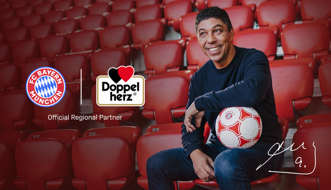 Un homme souriant est assis dans un stade vide, tenant un ballon de football. À côté de lui se trouvent les logos du FC Bayern Munich et de Doppelherz, indiquant un partenariat. L'homme porte un pull noir et un jean, et il dégage une atmosphère amicale et enthousiaste.
