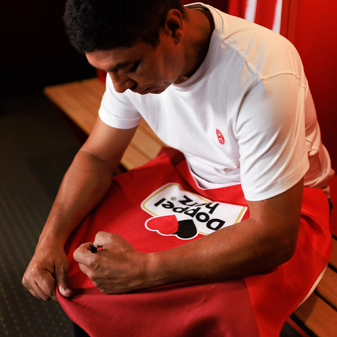 Un homme assis sur un banc dans une salle de sport, portant un t-shirt blanc, ajuste un vêtement rouge avec un logo coloré. Il semble concentré sur sa tâche. L'environnement est simple avec des murs et des sièges en bois.