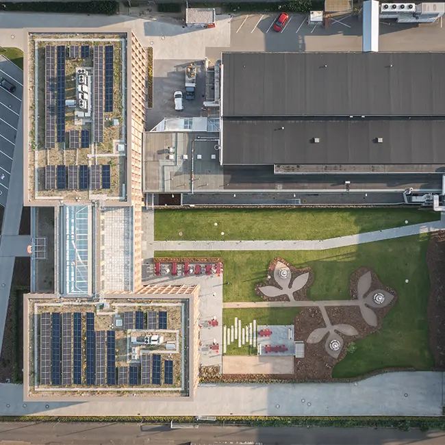 Vue aérienne d'un bâtiment moderne avec des toits végétalisés et des panneaux solaires. En dessous, un espace vert aménagé avec des chemins, des zones de repos et des motifs de jardin. Des voitures sont garées à proximité, et un bâtiment adjacent est visible sur le côté.