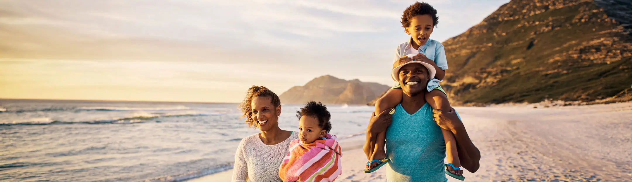 Une famille se tient sur une plage au coucher du soleil. Un homme porte un enfant sur ses épaules, tandis qu'une femme tient un autre enfant. Ils affichent tous des sourires, entourés de paysages côtiers.