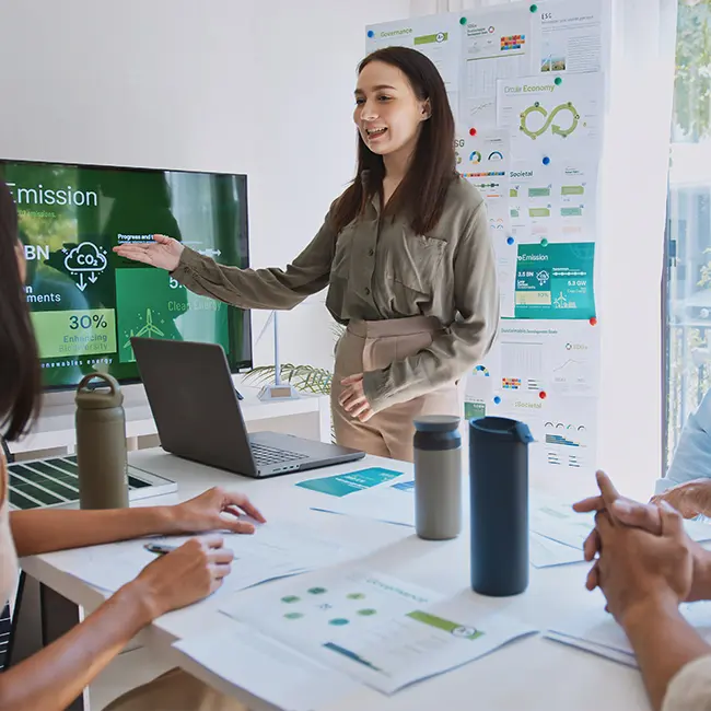 Une femme présente avec un écran de projection affichant des données sur les émissions de CO2. Elle se tient devant un ordinateur portable sur une table, entourée de documents et de bouteilles d'eau, tandis que d'autres personnes l'écoutent attentivement.