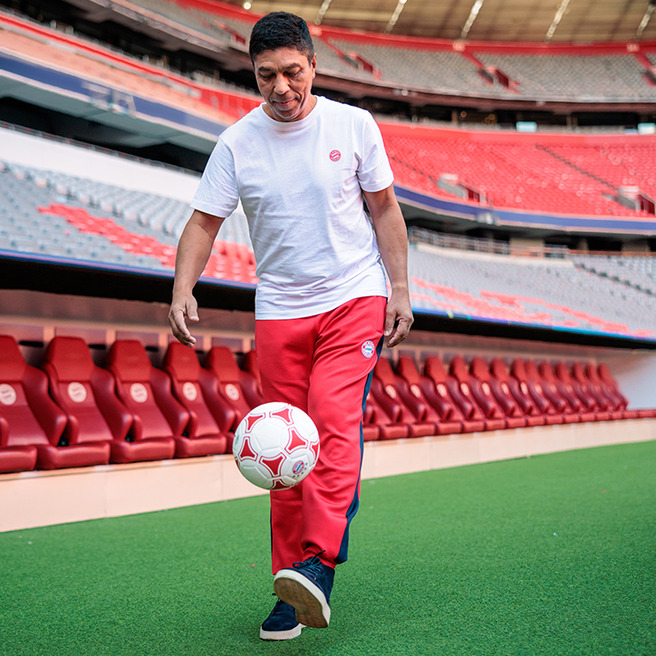 Un homme souriant porte un t-shirt blanc et un pantalon rouge, en train de jongler avec un ballon de football sur un terrain. En arrière-plan, on aperçoit des sièges rouges vides dans un stade.