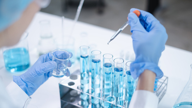Un scientifique en blouse de laboratoire et gants bleus utilise une pipette pour ajouter un liquide à des tubes à essai contenant un liquide bleu. Un bécher est aussi visible dans sa main. L'environnement est un laboratoire.