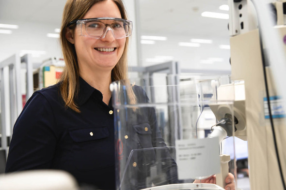 Femme souriante portant des lunettes de protection, se tenant près d'un équipement de laboratoire. L'environnement est moderne et bien éclairé, avec des étagères d'articles de laboratoire en arrière-plan.