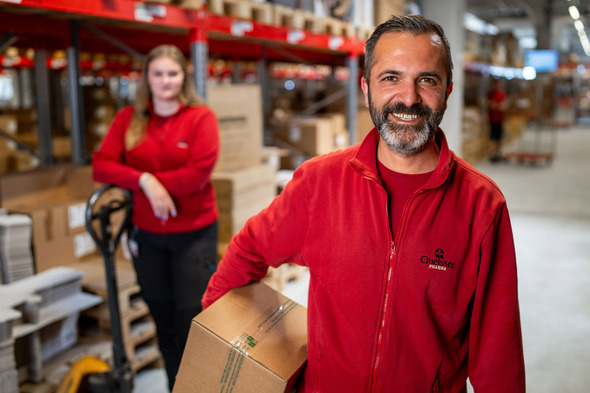 Un homme souriant portant une boîte se tient au premier plan, vêtu d'un manteau rouge. En arrière-plan, une femme en rouge se tient près d'un chariot élévateur, entourée de cartons dans un entrepôt. L'ambiance est professionnelle et dynamique.