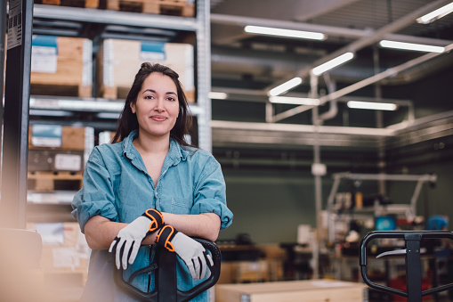 Femme souriante, habillée d'une chemise en denim, tenant un chariot élévateur dans un environnement de travail, entourée de caisses empilées et d'équipements industriels en arrière-plan.